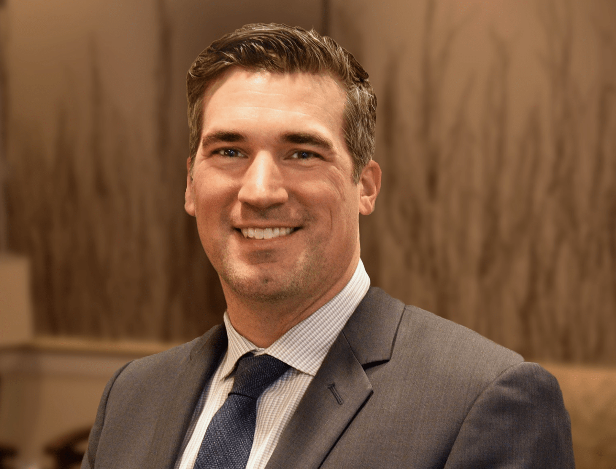 A man in a gray suit, light blue dress shirt, and dark blue tie smiles at the camera. The background is softly blurred with warm brown tones and abstract patterns.