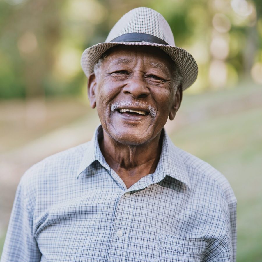 Portrait of a senior man looking at camera Smiling elderly man wearing a light-colored hat and a plaid shirt stands outdoors with blurred greenery in the background.