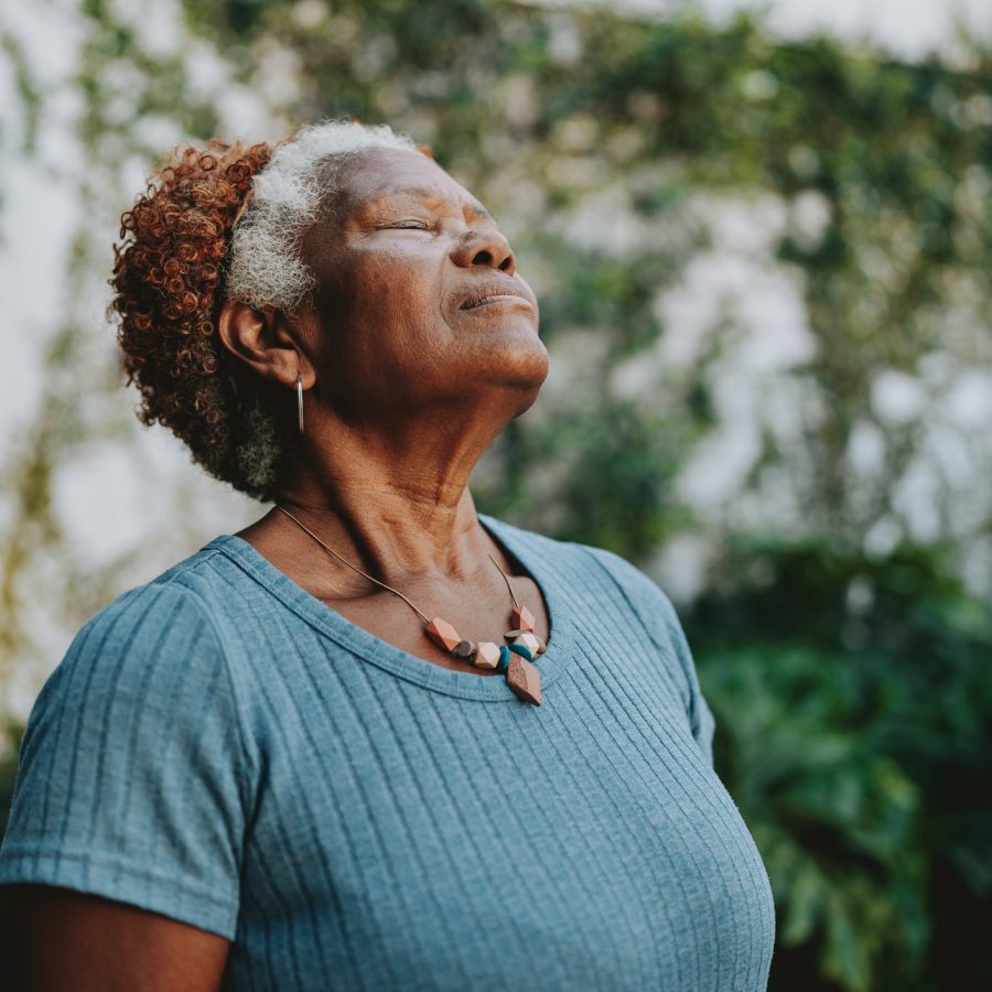 An older woman with short curly hair stands outdoors with her eyes closed, head tilted slightly up, and a peaceful expression. She wears a blue shirt and a beaded necklace, surrounded by greenery.