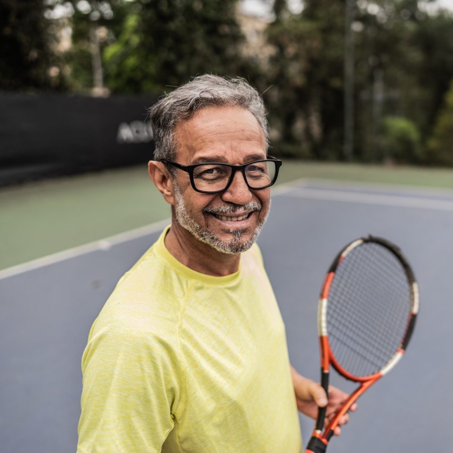 Portrait of a senior man in a tennis court Smiling man with glasses holds a tennis racket on an outdoor tennis court, wearing a yellow shirt. Trees and a fence are visible in the background.