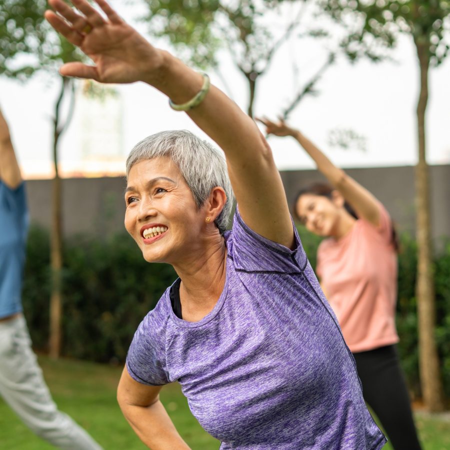 group of sportsperson dancing zumba outdoor Three people exercise outdoors, stretching with one arm raised. A smiling older woman in a purple shirt is in the foreground, with two others in the background. They are surrounded by greenery and trees.