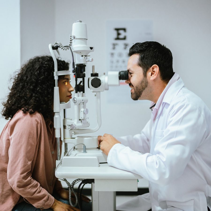 Young woman doing optical exam at medical clinic An eye doctor in a white coat examines a female patient's eyes using a slit lamp in a clinic. The patient sits with her chin on the device while the doctor looks through the microscope. An eye chart is visible in the background.