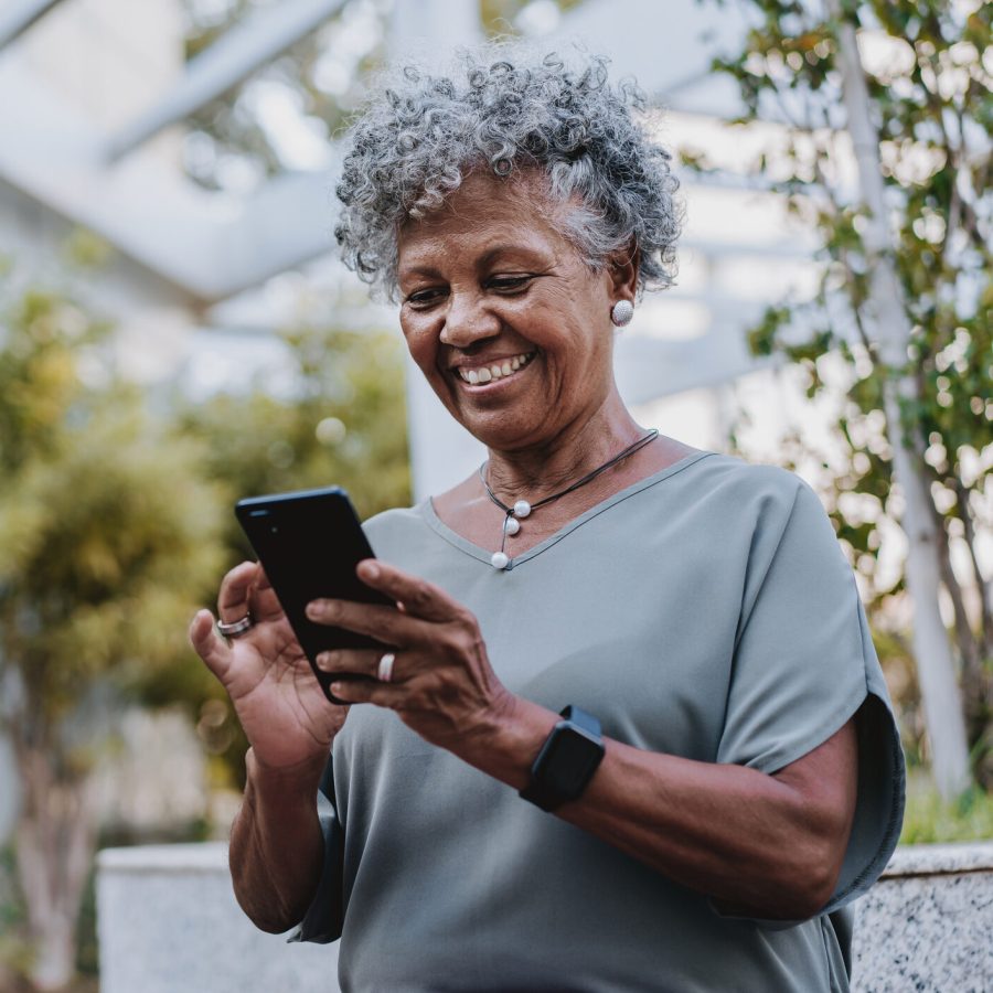 Portrait of a senior woman using smartphone An older woman with curly gray hair smiles while using a smartphone outdoors. She wears a gray blouse, a necklace, stud earrings, and a smartwatch, standing in a garden-like setting with greenery in the background.