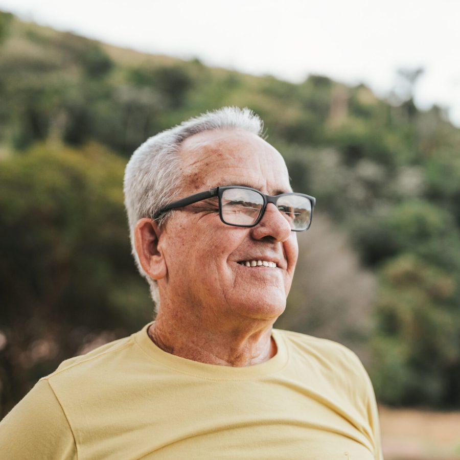 Portrait of a contemplative happy senior man An older man with short gray hair and glasses smiles while looking into the distance, standing outdoors with blurred green trees in the background. He is wearing a light yellow t-shirt.