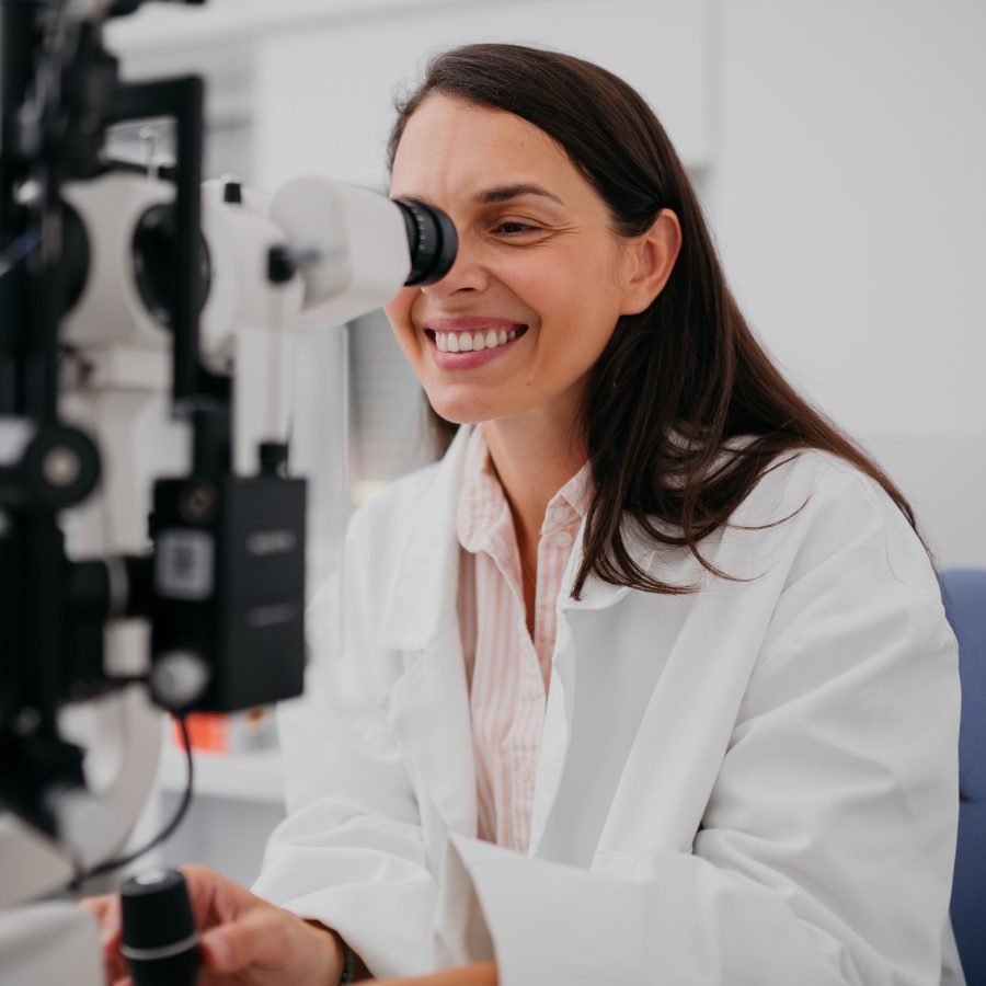 An eye exam A woman in a white lab coat smiles while looking into an eye examination device in a bright, modern medical office.