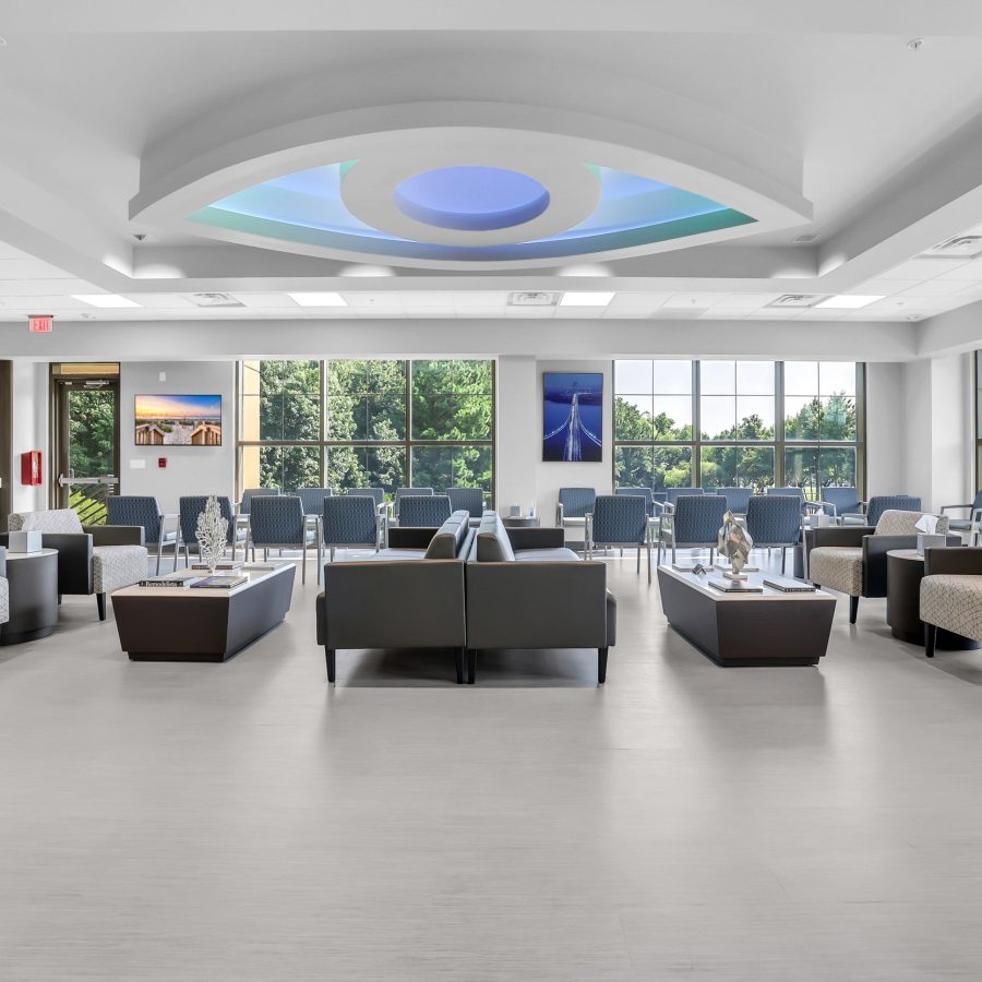 des_1668_1 A spacious, modern waiting room with rows of chairs and sofas, large windows letting in natural light, a coffee station on the left, and landscape photographs on the walls. The ceiling features a blue circular design.